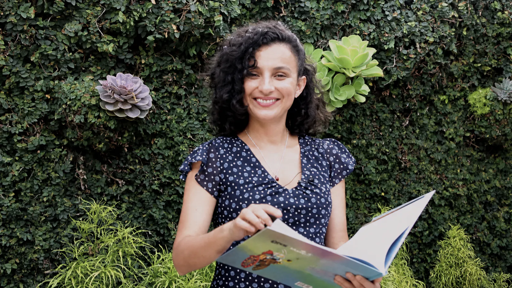 Hazel Hernández Astorga sonriendo, con un libro abierto frente a un jardín vertical